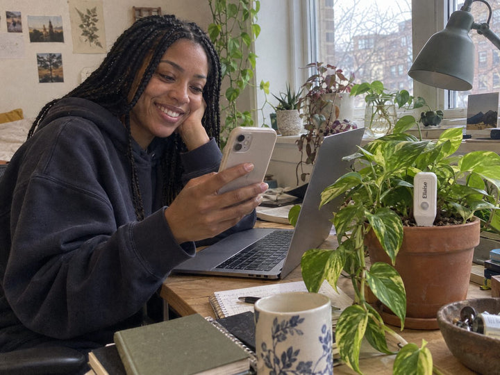Young woman at a desk with a pothos and an Ellaise sensor, phone in her hand.
